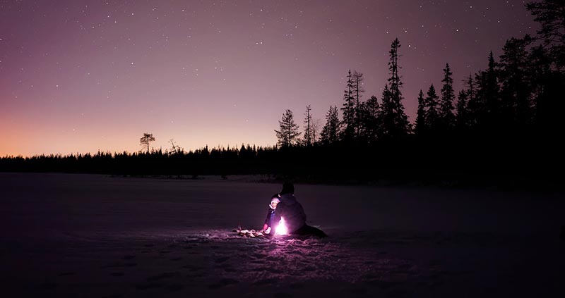 Person sitter ved et lite bål i vinterlandskap under stjernehimmel