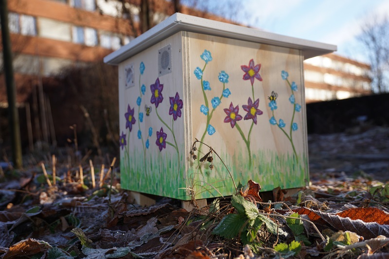 Humlekasse dekorert med blomstermotiver, plassert utendørs i frostrikt gress