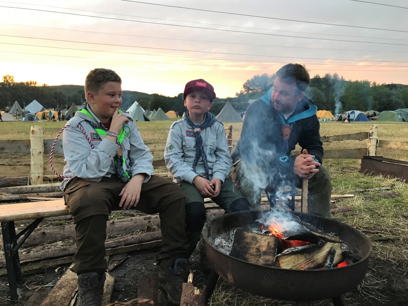 To speidergupper og en leder sitter rundt bålet på en leir. 