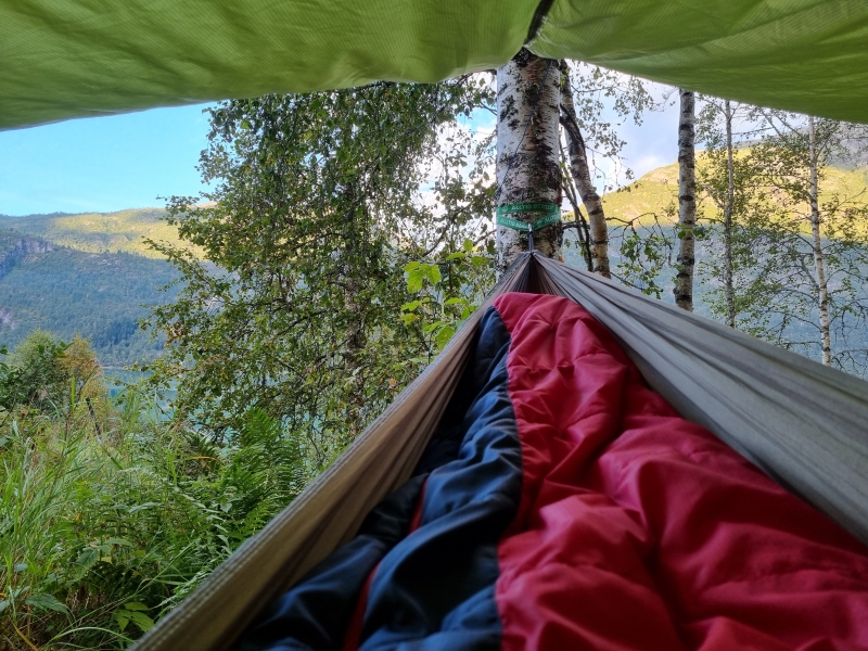 Hengekøye med sovepose under presenning i skogen med fjord og fjell i bakgrunnen