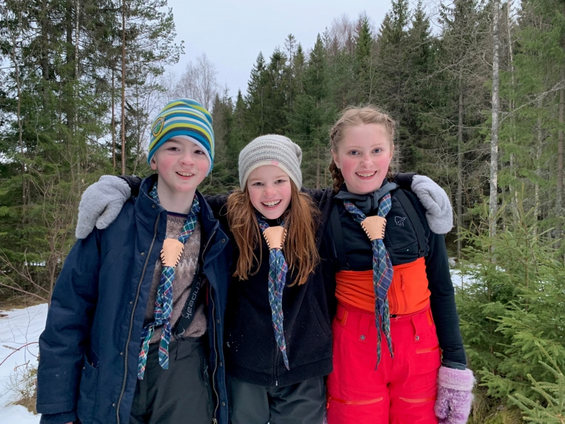 Tre speidere smiler og holder rundt hverandre i skogen om vinteren