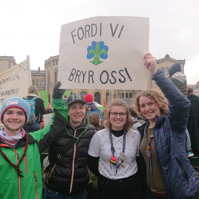 Fire unge speidere foran Stortinget med plakat der det står "Fordi vi bryr oss!"