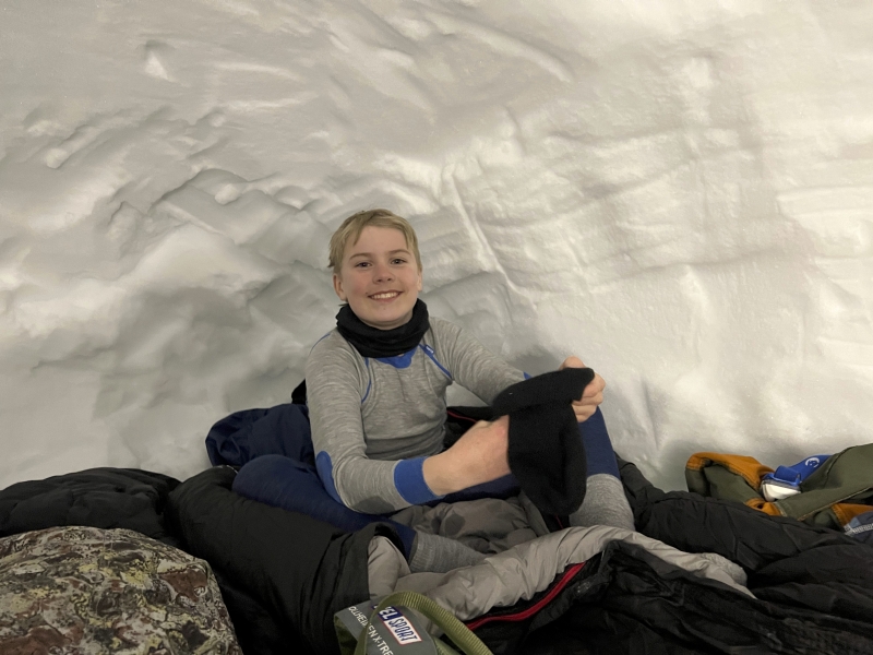 Ung speider sitter smilende i sovepose inne i en snøhule